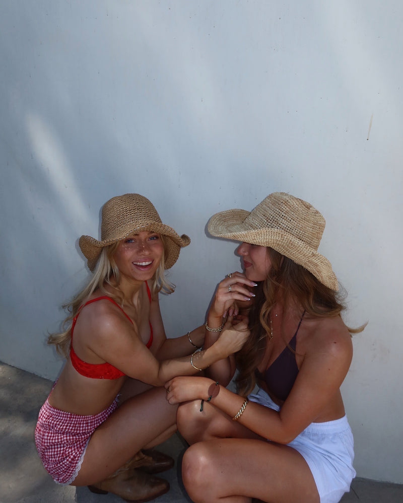 Two girls wearing bikinis and cowboy hats crouching down and smiling, looking very tanned