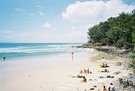 Noosa beach with people sunbathing and trees just off the rocks over the water